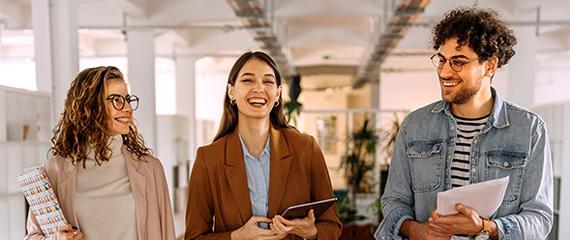 Tre persone camminano in un moderno spazio di coworking luminoso, con documenti e dispositivi in mano, tra colonne bianche e aree verdi interne. | Crédit Agricole Tre persone camminano in un moderno spazio di coworking luminoso, con documenti e dispositivi in mano, tra colonne bianche e aree verdi interne. | Crédit Agricole