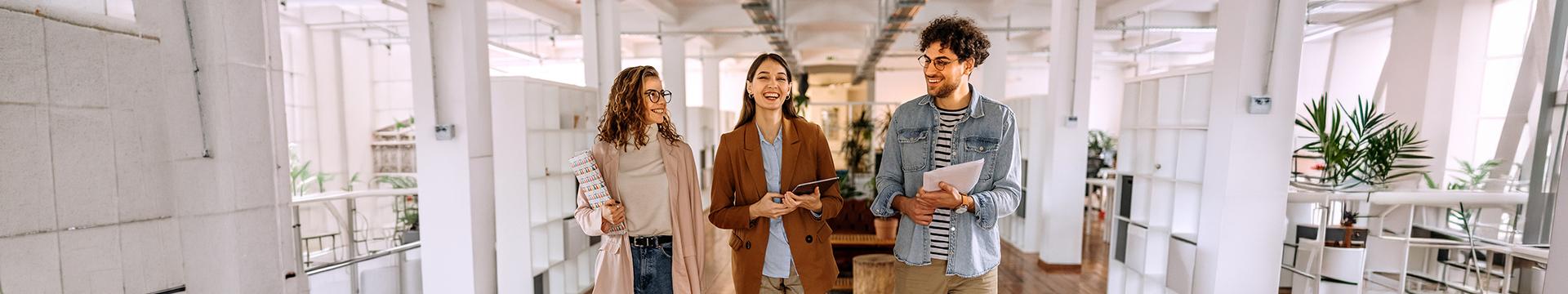 Tre persone camminano in un moderno spazio di coworking luminoso, con documenti e dispositivi in mano, tra colonne bianche e aree verdi interne. | Crédit Agricole Tre persone camminano in un moderno spazio di coworking luminoso, con documenti e dispositivi in mano, tra colonne bianche e aree verdi interne. | Crédit Agricole
