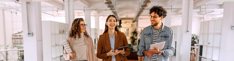 Tre persone camminano in un moderno spazio di coworking luminoso, con documenti e dispositivi in mano, tra colonne bianche e aree verdi interne. | Crédit Agricole Tre persone camminano in un moderno spazio di coworking luminoso, con documenti e dispositivi in mano, tra colonne bianche e aree verdi interne. | Crédit Agricole
