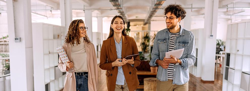 Tre persone camminano in un moderno spazio di coworking luminoso, con documenti e dispositivi in mano, tra colonne bianche e aree verdi interne. | Crédit Agricole Tre persone camminano in un moderno spazio di coworking luminoso, con documenti e dispositivi in mano, tra colonne bianche e aree verdi interne. | Crédit Agricole