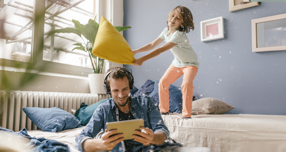 papà che guarda il tablet con le cuffie e bambina dietro di lui sul divano che sta per colpirlo con un cuscino | Crédit Agricole papà che guarda il tablet con le cuffie e bambina dietro di lui sul divano che sta per colpirlo con un cuscino | Crédit Agricole